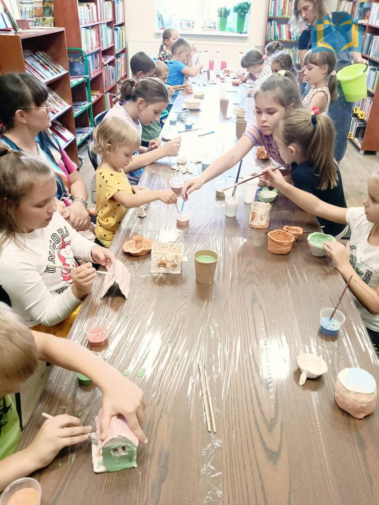 Zdjęcie: W bibliotece dzieci malują swoje wyroby z gliny. Siedzą przy długim, brązowym stole, na którym znajdują się kubeczki z farbami.